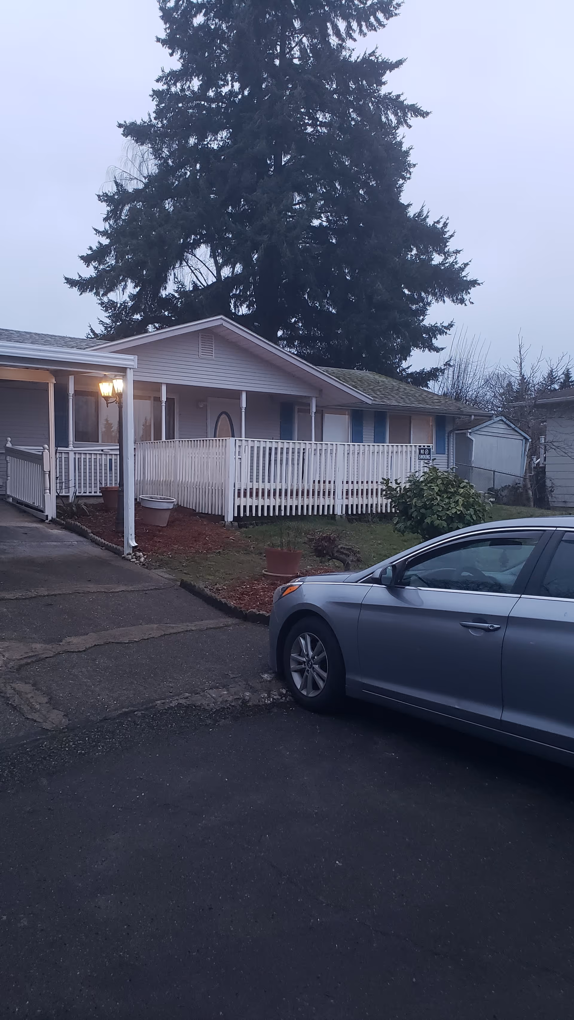 Exterior view of a single-story residential building with a white railing and a ramp leading to the entrance. A large evergreen tree is visible behind the building. A silver car is parked in the driveway in front of the building. The sky is overcast and the outdoor light near the entrance is on.