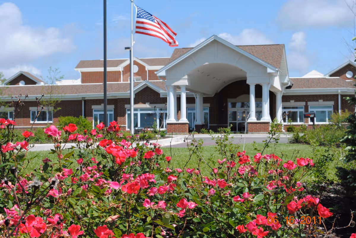 Front exterior view of a brick building with a covered entrance supported by white columns, an American flag on a flagpole, and vibrant pink flowers in the foreground under a partly cloudy blue sky.