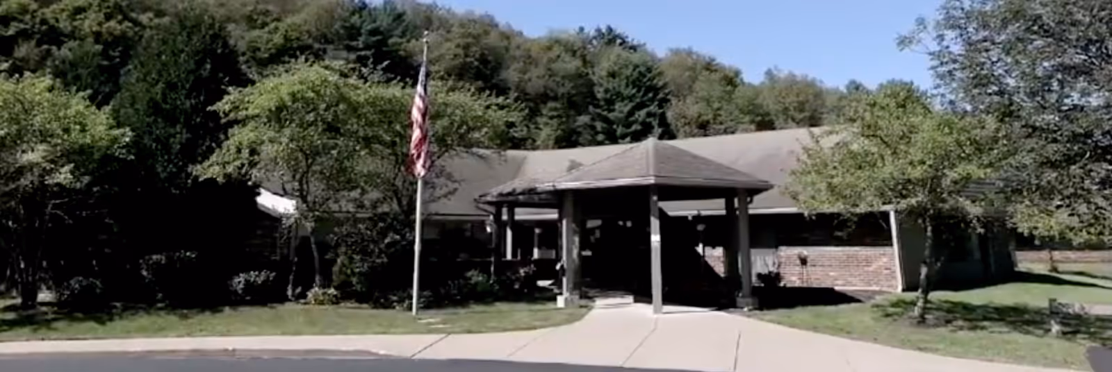Exterior view of a single-story building with a covered entrance, surrounded by trees and greenery under a clear blue sky. An American flag is displayed on a flagpole near the entrance.