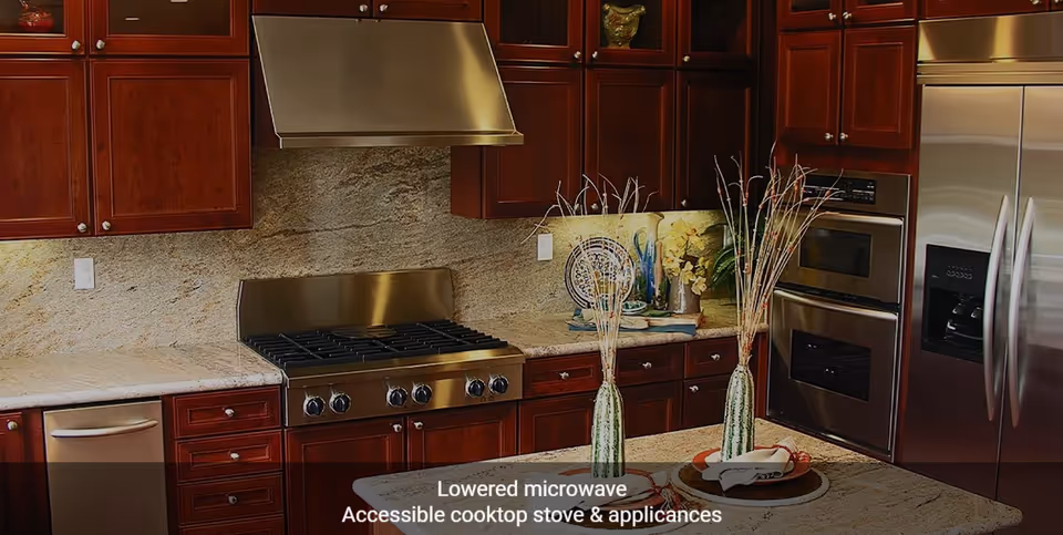 Modern kitchen with dark wood cabinets, stainless steel appliances including a lowered microwave, accessible cooktop stove, double oven, dishwasher, and refrigerator. The kitchen island has two decorative vases with tall branches and place settings.
