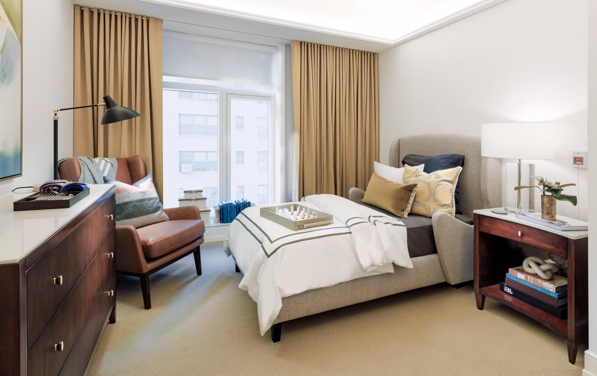 A bright and cozy bedroom featuring a beige upholstered bed with white and beige bedding, a tray with a chess set on the bed, a wooden nightstand with a lamp and decorative items, a brown leather armchair with a pillow and throw blanket, a wooden dresser with a tray on top, and large windows with beige curtains letting in natural light.