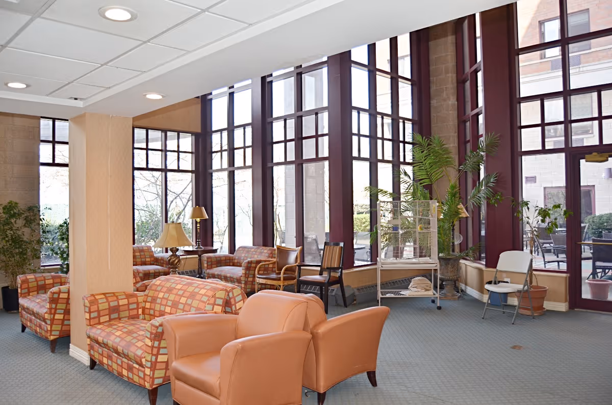 A bright and spacious living room area with large floor-to-ceiling windows letting in natural light. The room is furnished with patterned armchairs, leather chairs, wooden chairs, and small tables with lamps. There are several potted plants and a birdcage near the windows. Outside the windows, patio furniture is visible.