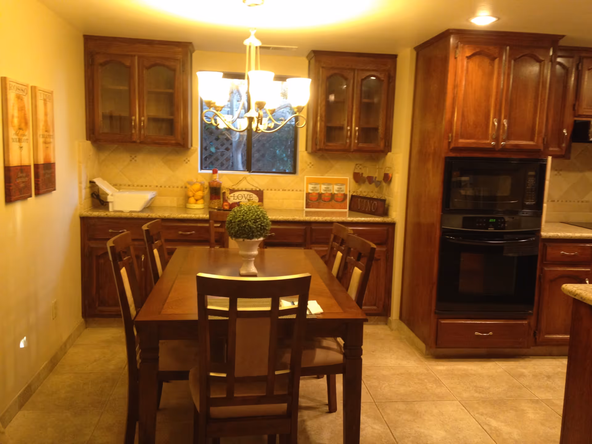 A warm kitchen and dining area with wooden cabinets and a wooden dining table surrounded by six chairs. A chandelier hangs above the table, which has a small green plant centerpiece. The kitchen features a built-in oven, microwave, and a window above the countertop. Decorative items and framed pictures adorn the walls and countertops.