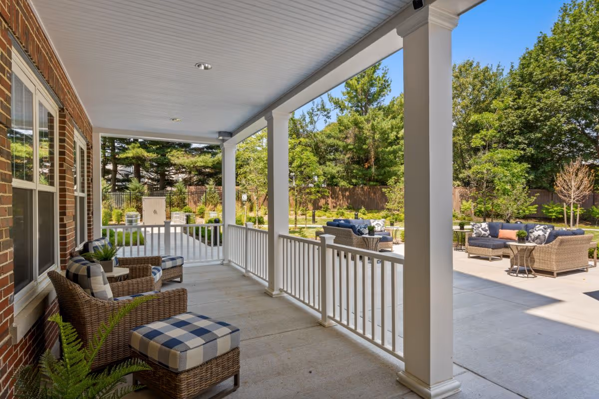 Covered porch area with wicker chairs and ottomans featuring blue and white checkered cushions, overlooking an outdoor patio with wicker sofas and tables surrounded by greenery and trees under a clear blue sky.
