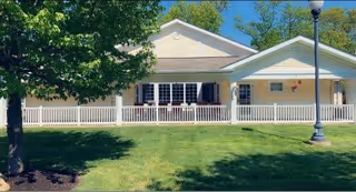 Single-story building with a white railing porch, beige siding, and a well-maintained green lawn in front. There is a tree on the left side and a streetlamp on the right side under a clear blue sky.