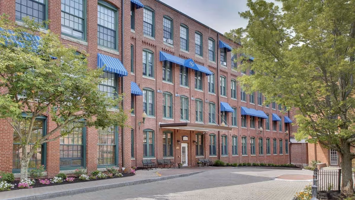 Exterior view of a multi-story brick building with green-trimmed windows and blue striped awnings. There are trees and flower beds along the sidewalk, and benches near the entrance.