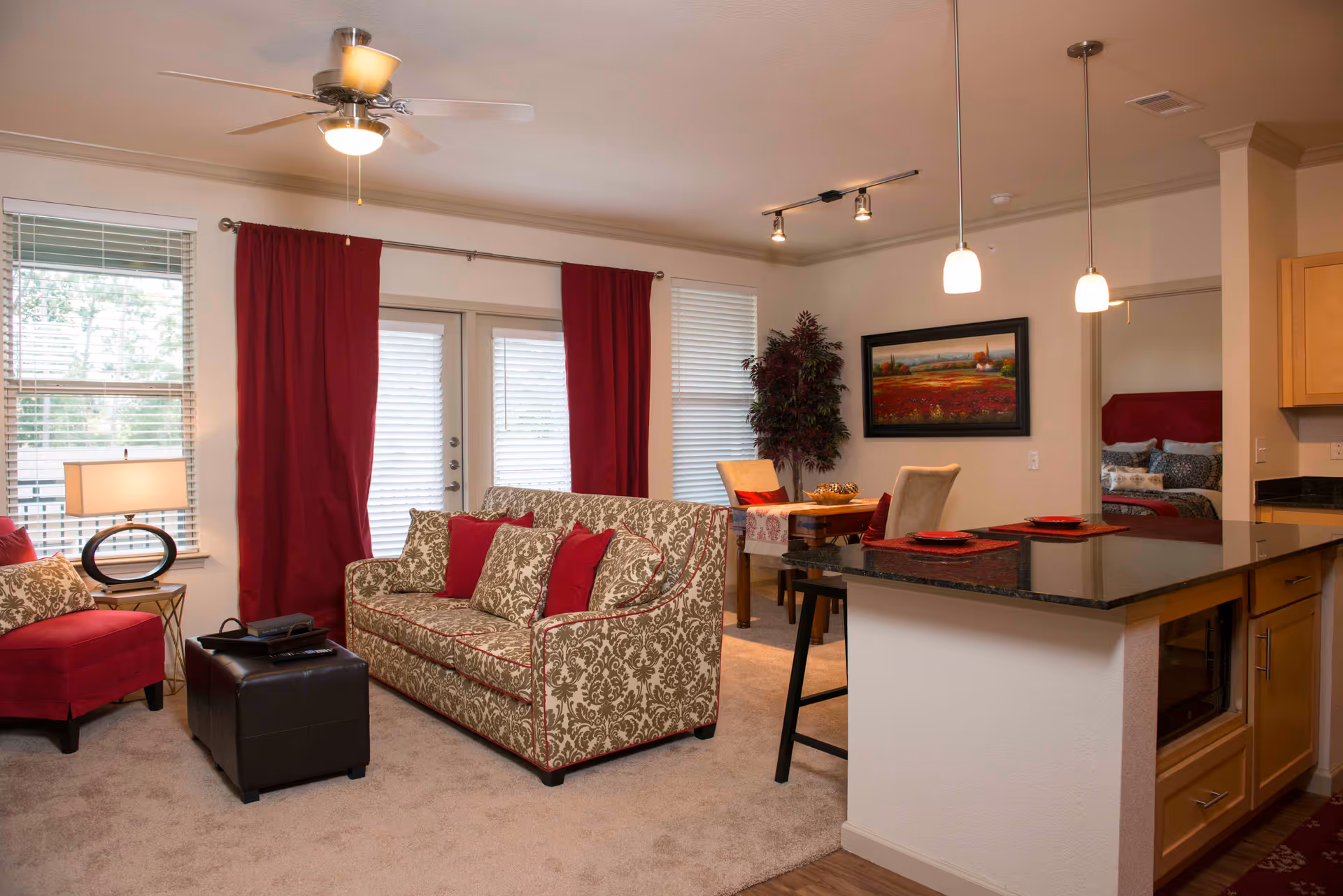 A cozy living room area with a patterned sofa and red cushions, a red armchair, a black ottoman, and a side table with a lamp. The room has large windows with red curtains and blinds, a ceiling fan with a light, and a small dining table with chairs near a framed landscape painting. The kitchen area with a granite countertop and pendant lights is visible on the right, and a bedroom with a red headboard is partially visible through an open doorway.