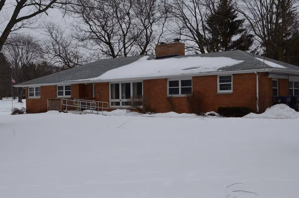 Single-story brick building with a gray roof partially covered in snow, surrounded by snow-covered ground and leafless trees in the background. There is a metal ramp leading to the entrance and an American flag visible on the left side.