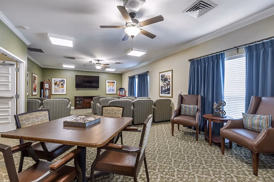 A cozy living room area in a senior living facility with multiple armchairs arranged facing a wall-mounted flat screen TV. The room features a wooden table with chairs in the foreground, patterned carpet, ceiling fans with lights, framed artwork on the walls, and large windows with blue curtains.