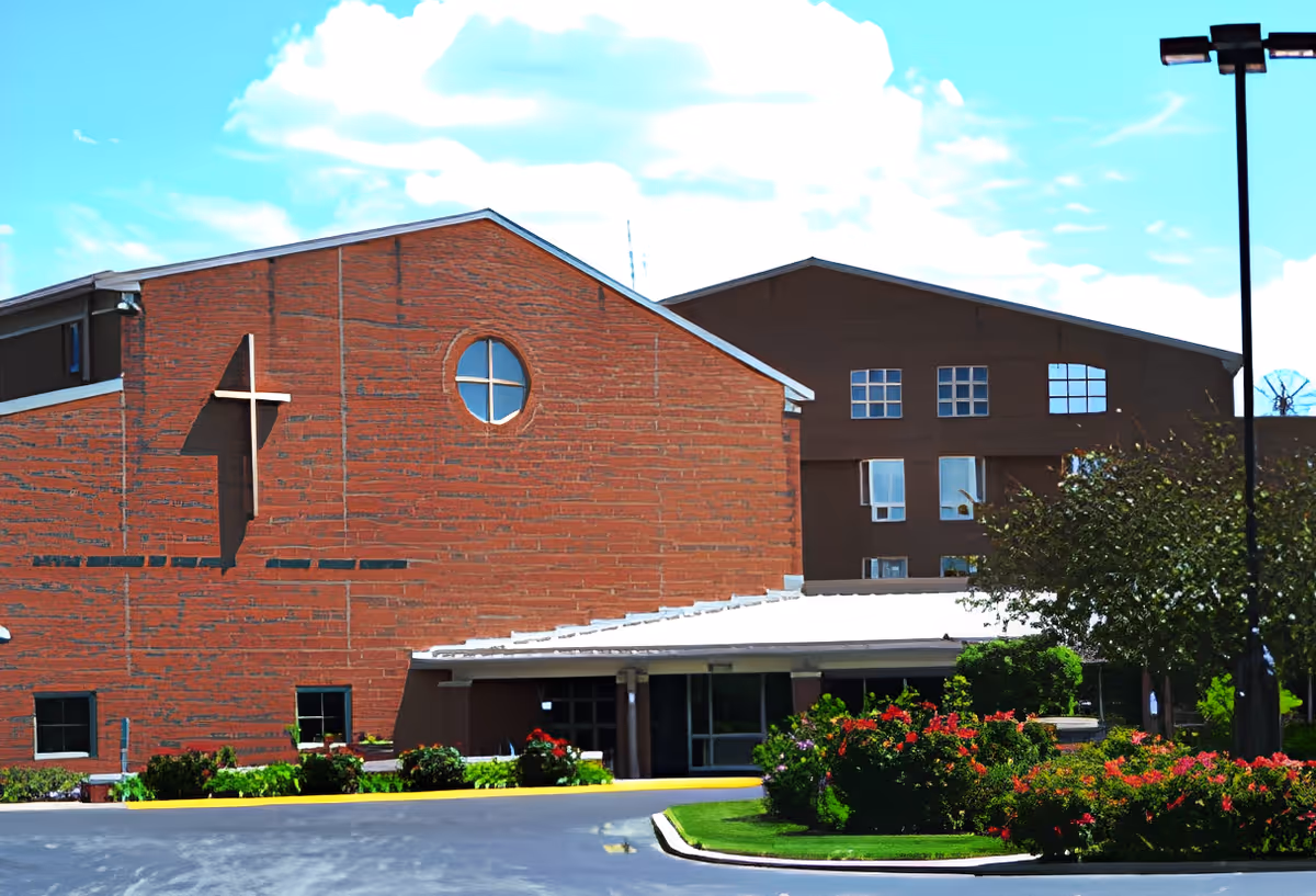 Exterior view of the Jeanne Jugan Center building featuring a brick facade with a cross and circular window, surrounded by landscaped bushes and flowers under a partly cloudy sky.
