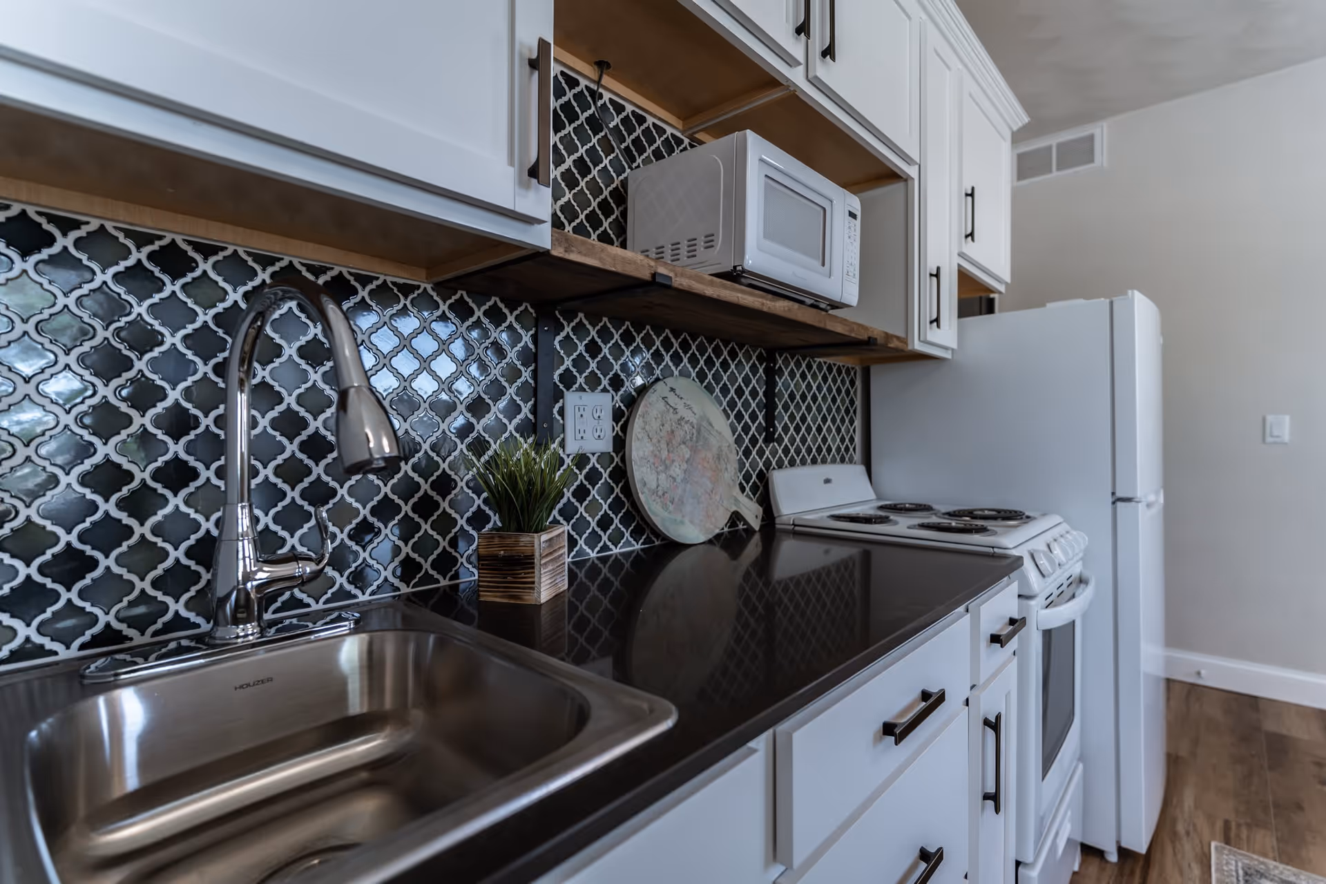 Modern kitchen interior with white cabinets, a stainless steel sink with a chrome faucet, a black countertop, a white microwave on a wooden shelf, a white electric stove, and a white refrigerator. The backsplash features a black and white patterned tile design.