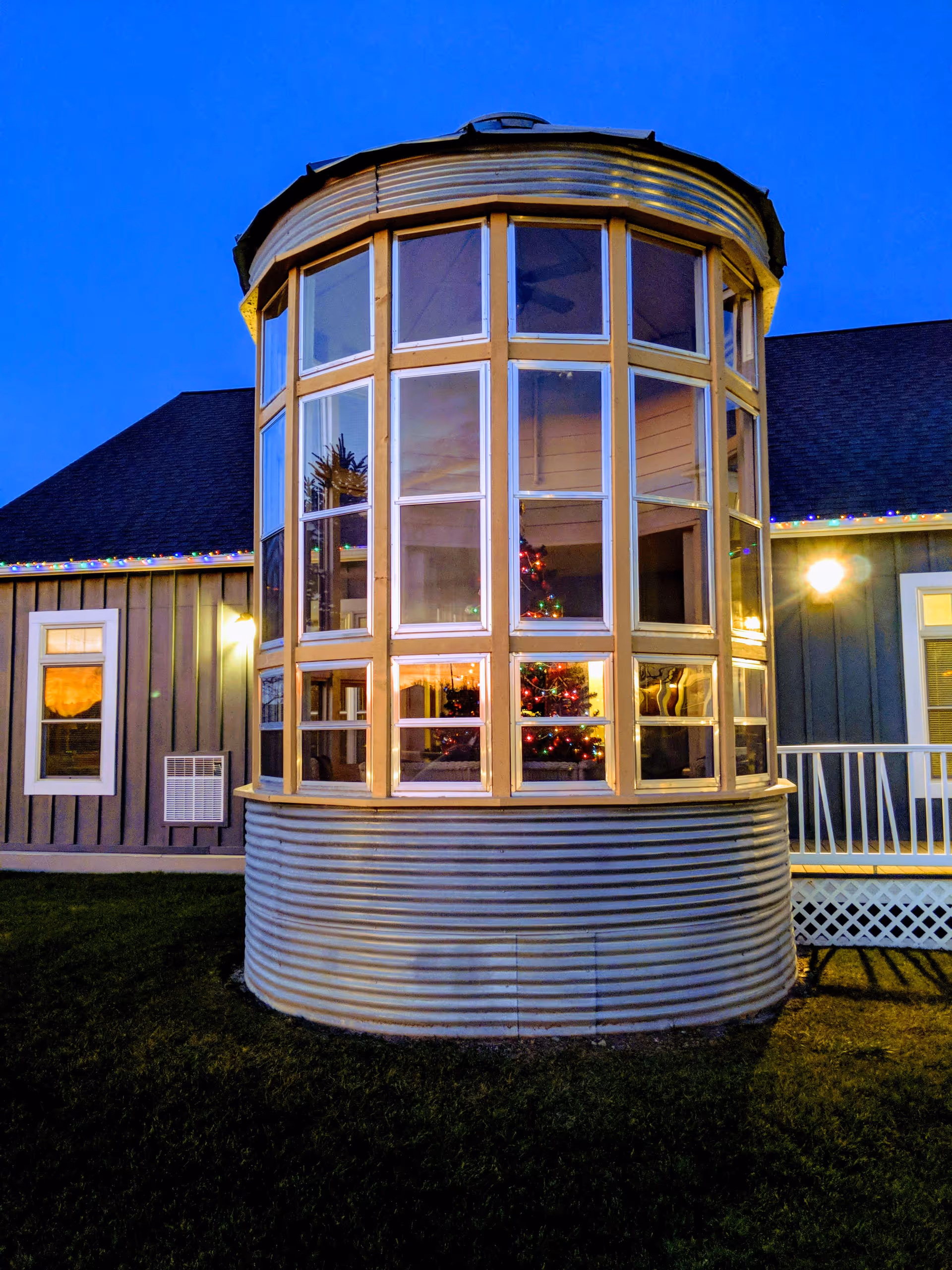 Exterior view of a building at dusk with a large, round, multi-pane window structure. Inside the window, a decorated Christmas tree with lights is visible. The building has gray siding, white trim, and a porch with white railing. The sky is clear and blue.
