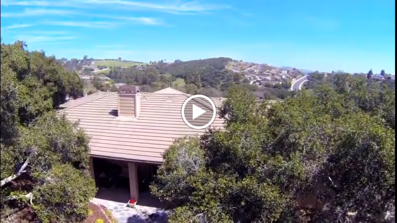 Aerial view of a senior living facility building with a tiled roof surrounded by trees and greenery, with hills and a residential area visible in the background under a clear blue sky.