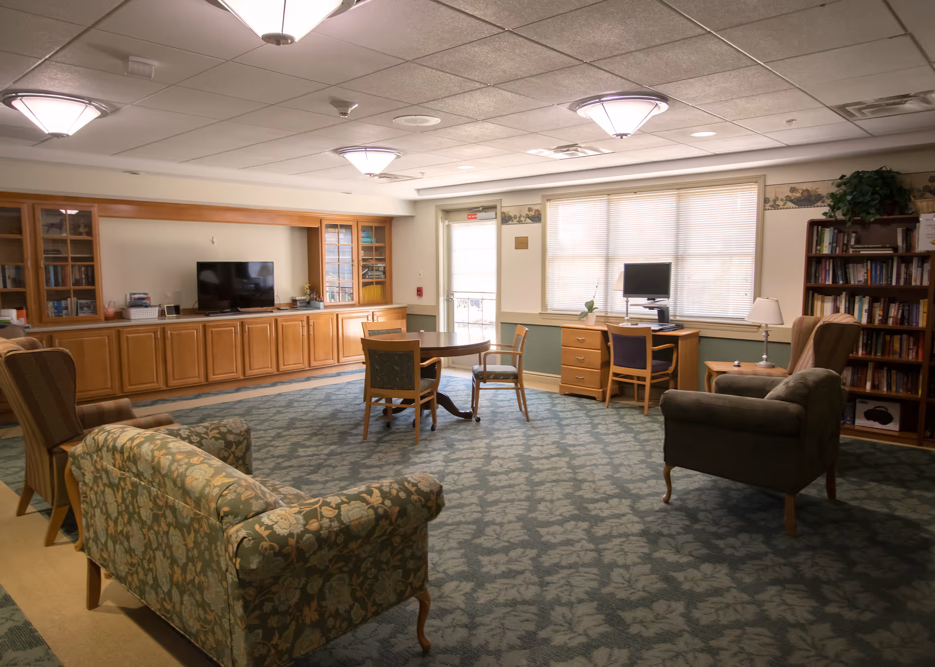 A cozy living room area in a senior living facility with patterned armchairs and a sofa, a round table with chairs in the center, a wooden cabinet with a TV, a desk with a computer, bookshelves filled with books, and large windows letting in natural light.