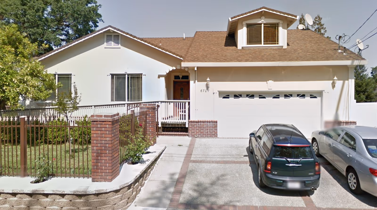 Front exterior view of a single-story residential building with a brown shingle roof, white walls, a garage door, and a small porch with a wooden front door. There are two cars parked in the driveway, a green Mini Cooper and a silver sedan. The property is enclosed by a metal fence with brick pillars, and there are trees and shrubs in the yard.