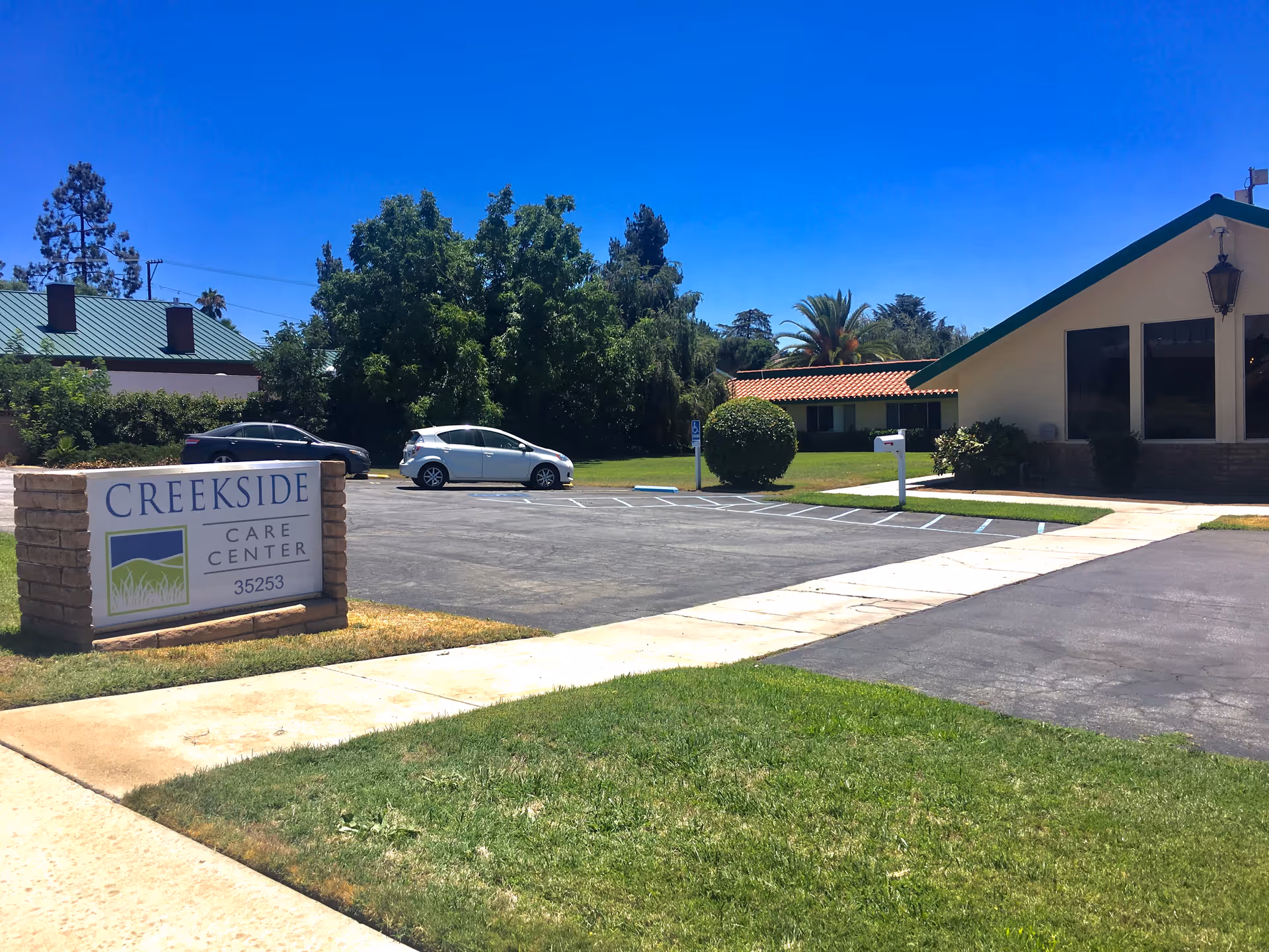 Exterior view of Creekside Care Center showing a parking lot with two cars, a sign with the facility name and address, green grass, trees, and a building with a green roof under a clear blue sky.