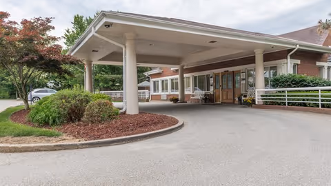 Entrance of a brick building with a covered driveway supported by large white columns, landscaped with bushes and trees, and a clear sky overhead.