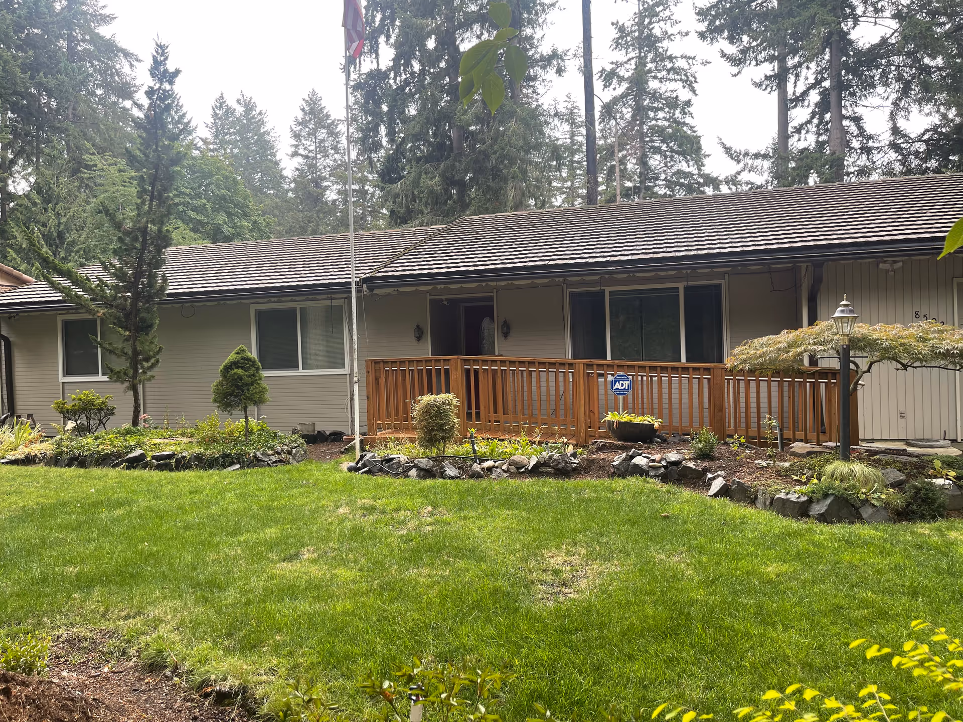 Single-story residential building with a wooden ramp leading to the entrance, surrounded by a well-maintained garden with green grass, shrubs, and trees. A flagpole is visible in front of the building, and tall trees are in the background.