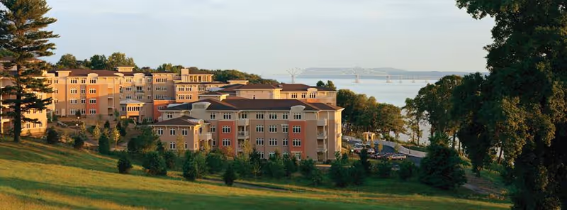 Wide exterior view of Kendal On Hudson facility buildings situated on a grassy hillside with trees around. In the background, a large body of water and a distant bridge are visible under a clear sky.