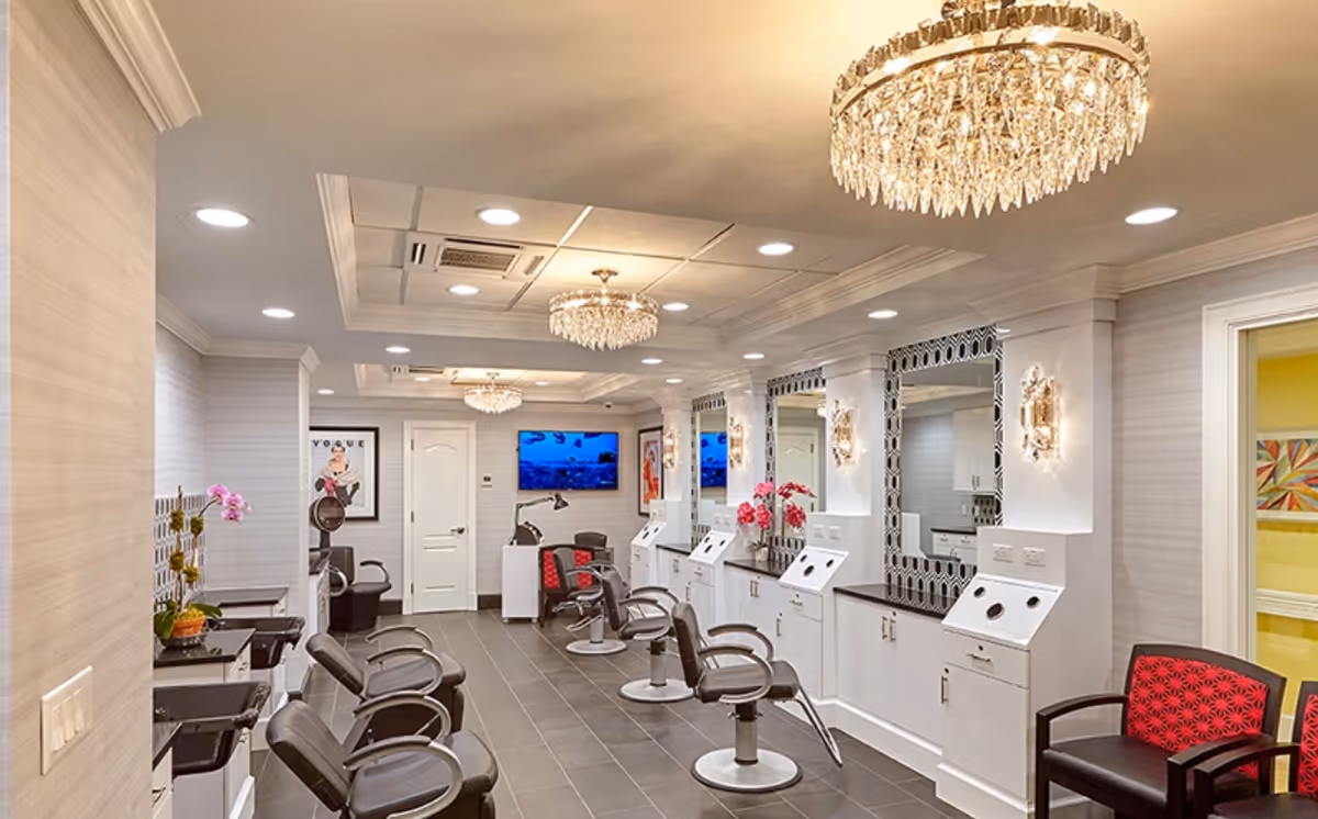 Interior view of a stylish salon area in an assisted living facility featuring multiple black salon chairs in front of large mirrors with white cabinetry, elegant chandeliers hanging from the ceiling, and a television mounted on the far wall. The space is well-lit with recessed lighting and decorated with framed artwork and flowers.