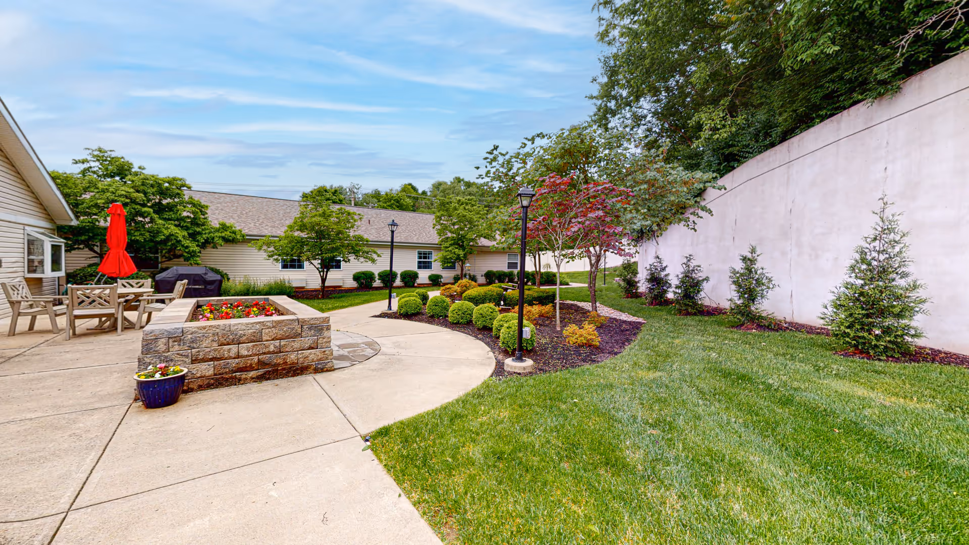 Outdoor courtyard area with a concrete walkway, green grass, and landscaped bushes and trees. There is a raised stone planter with flowers, patio chairs, a table with a red umbrella, and a barbecue grill. The area is surrounded by buildings and a tall white wall.