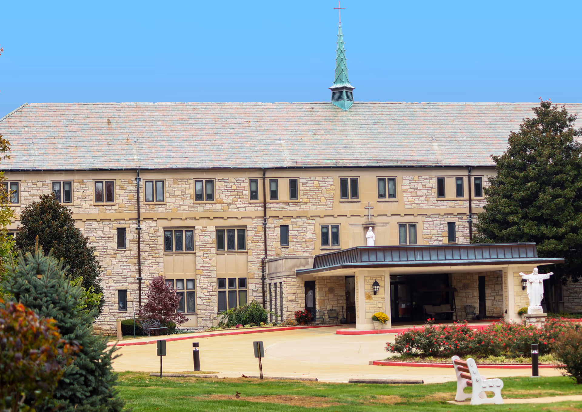 Exterior view of a large stone building with multiple windows and a peaked roof with a cross on top. The entrance has a covered driveway with columns, surrounded by landscaped bushes and trees. A white statue is visible near the entrance, and a white bench is placed on the grass in the foreground.