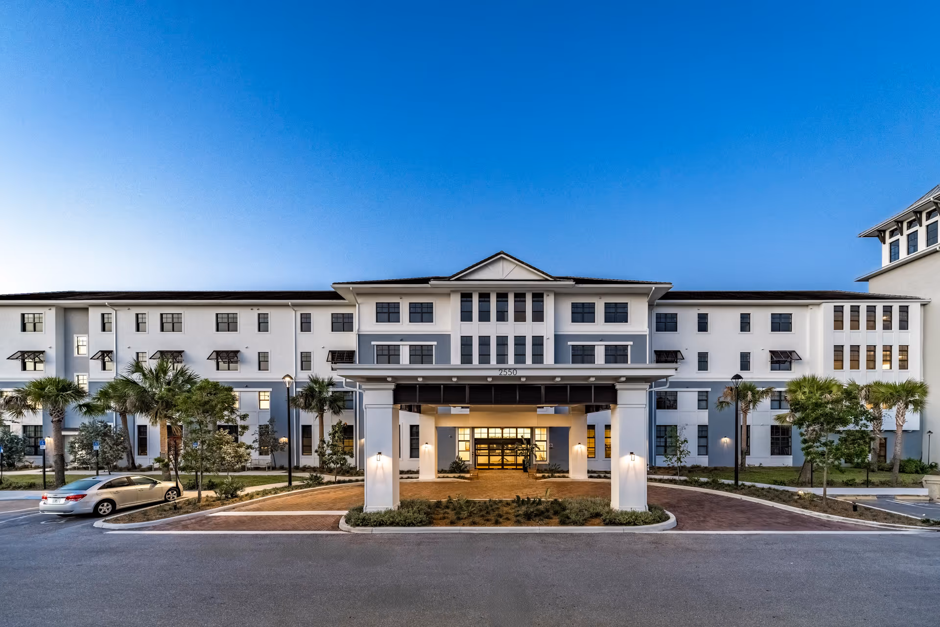 Front exterior view of a large, modern senior living facility building with a covered entrance, palm trees, and a car parked in front under a clear blue sky.