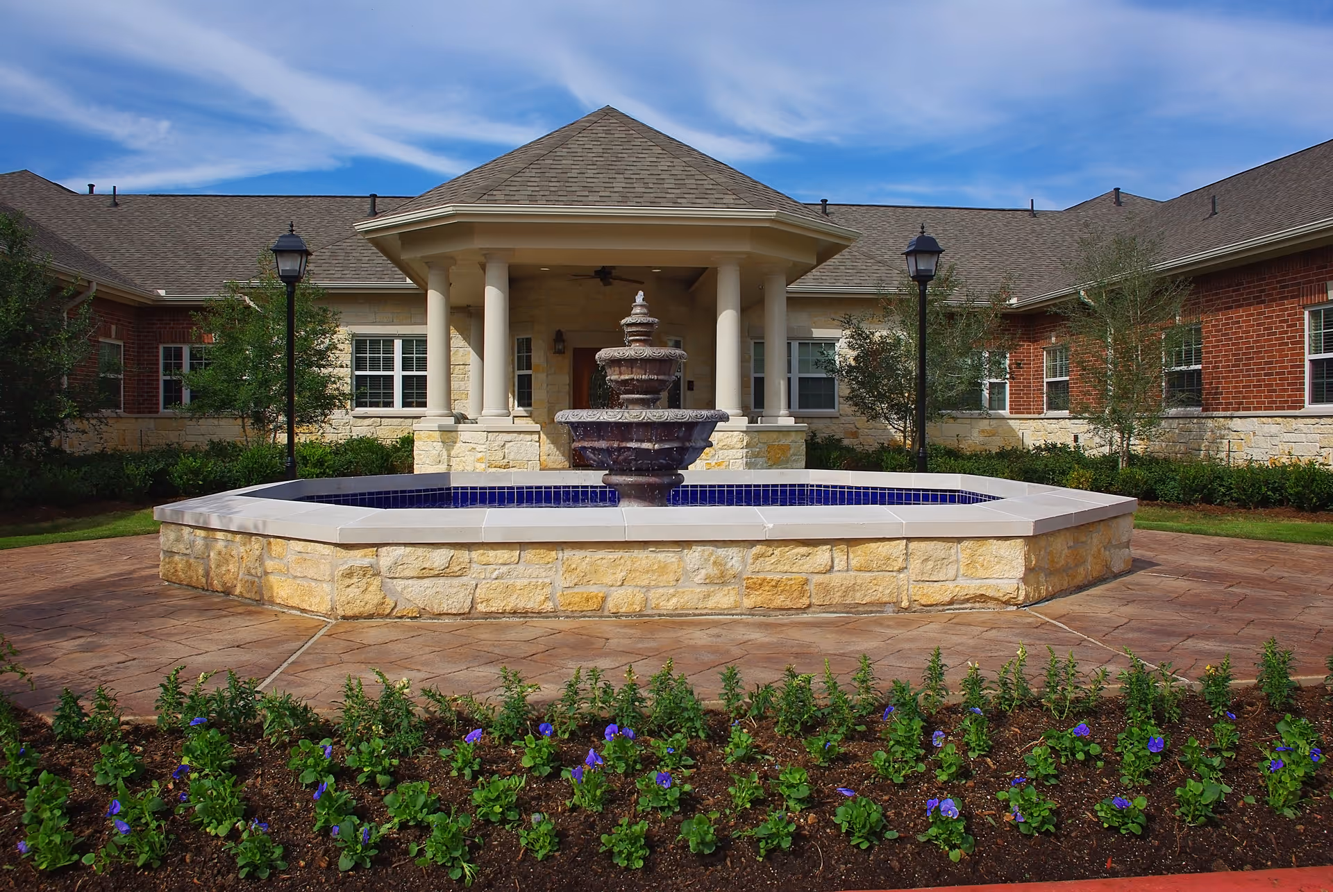 Outdoor view of The Heritage of Meyerland facility featuring a stone fountain in the center surrounded by a paved walkway, flower beds with purple flowers in the foreground, and a building with brick and stone exterior walls and a covered entrance with columns in the background under a blue sky with some clouds.