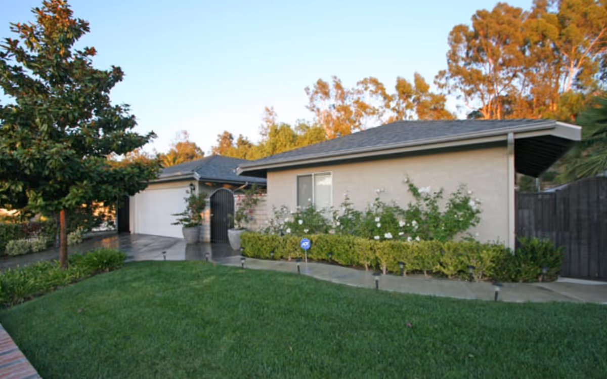 Exterior view of a single-story residential building with a well-maintained green lawn, a tree on the left, and bushes with white flowers along the front wall. The building has a gray roof and beige walls, with a garage door and a black gate visible.