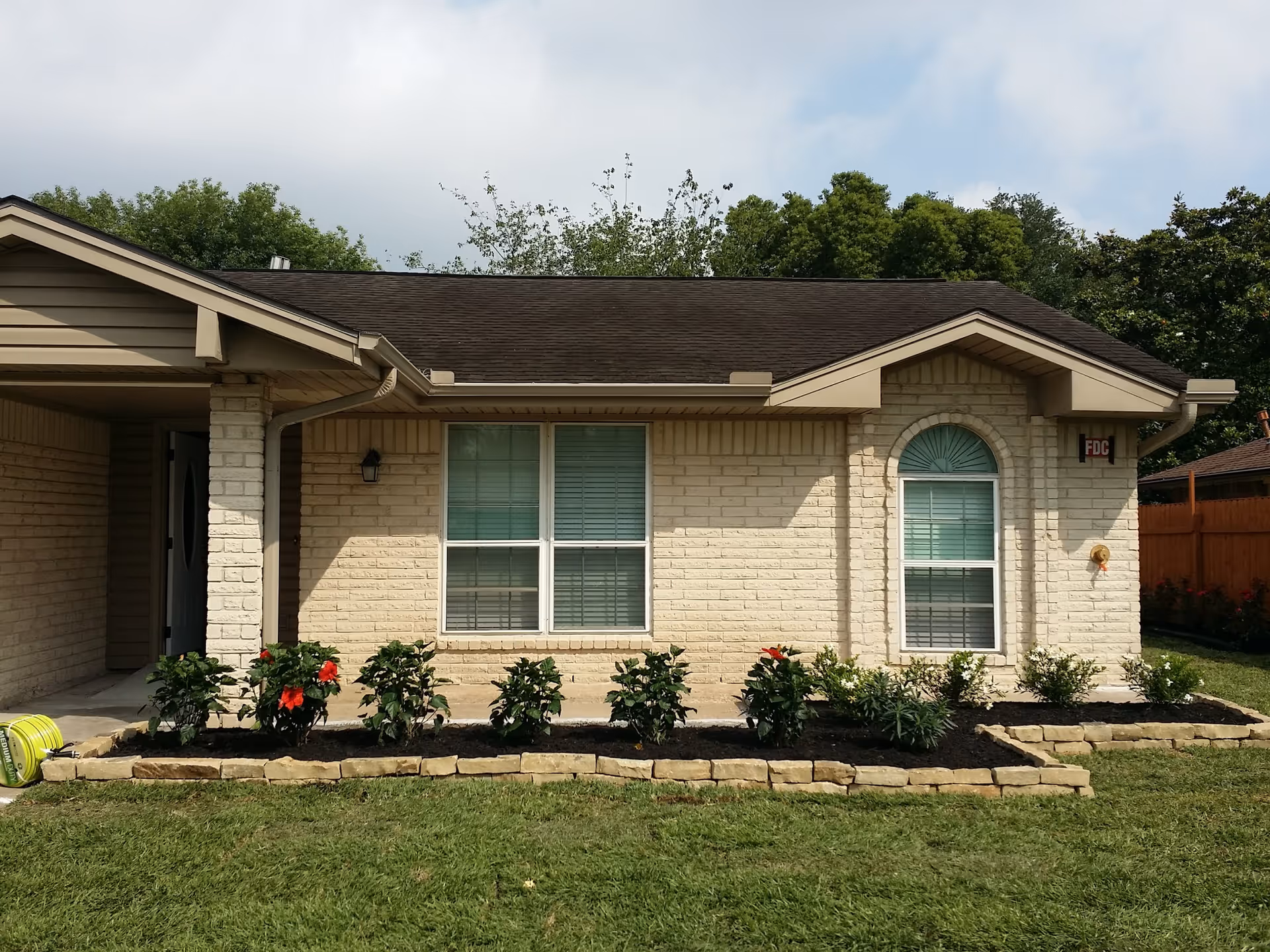 Exterior view of a single-story beige brick building with two windows, a small garden bed with flowering plants in front, and a grassy lawn. The sky is partly cloudy and there are trees in the background.