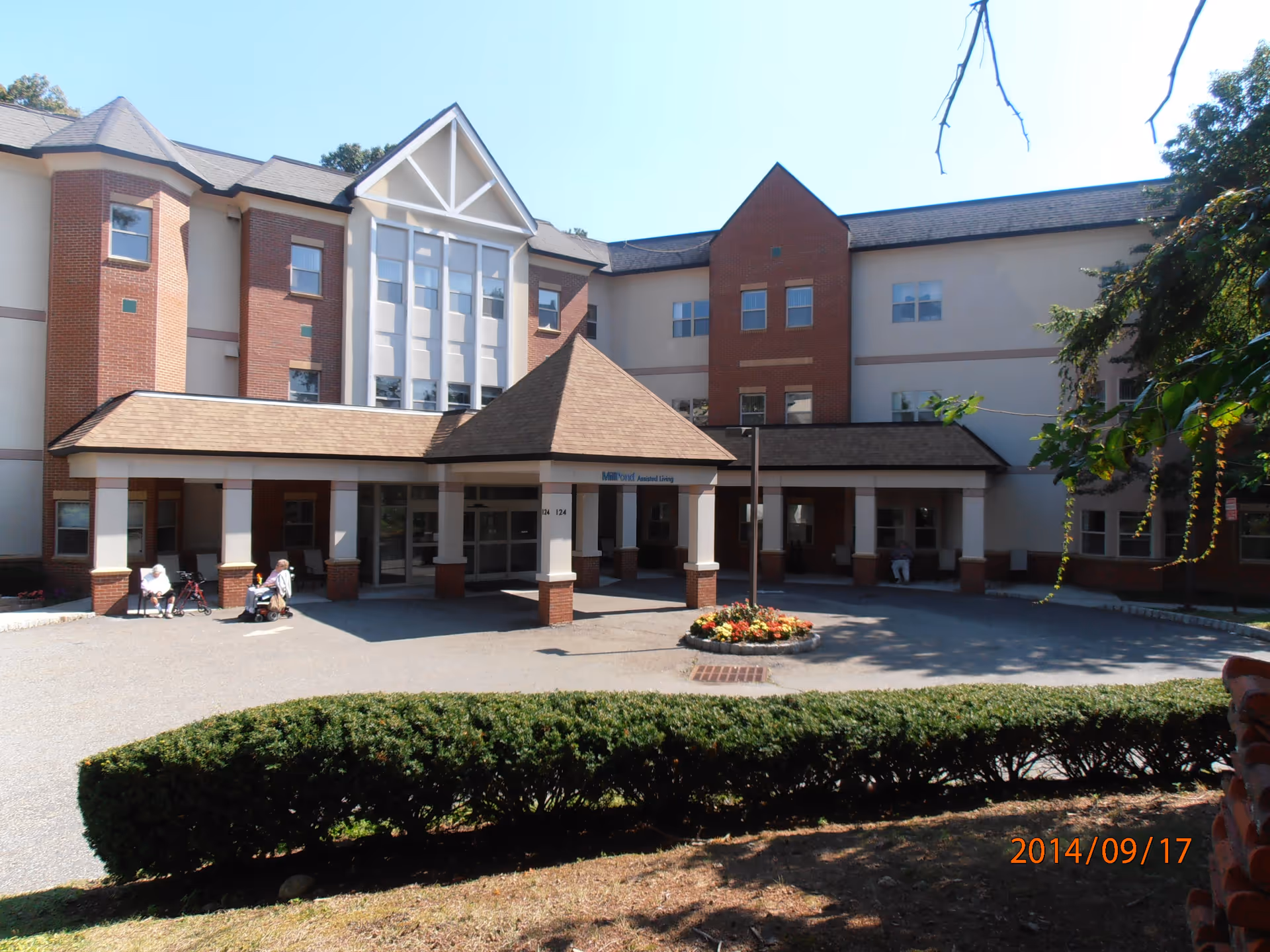 Exterior view of a multi-story assisted living facility with a covered entrance, brick and beige walls, and several windows. There are a few elderly people sitting near the entrance, some with walkers. A small circular flower bed is in front of the entrance, and trimmed bushes line the foreground.