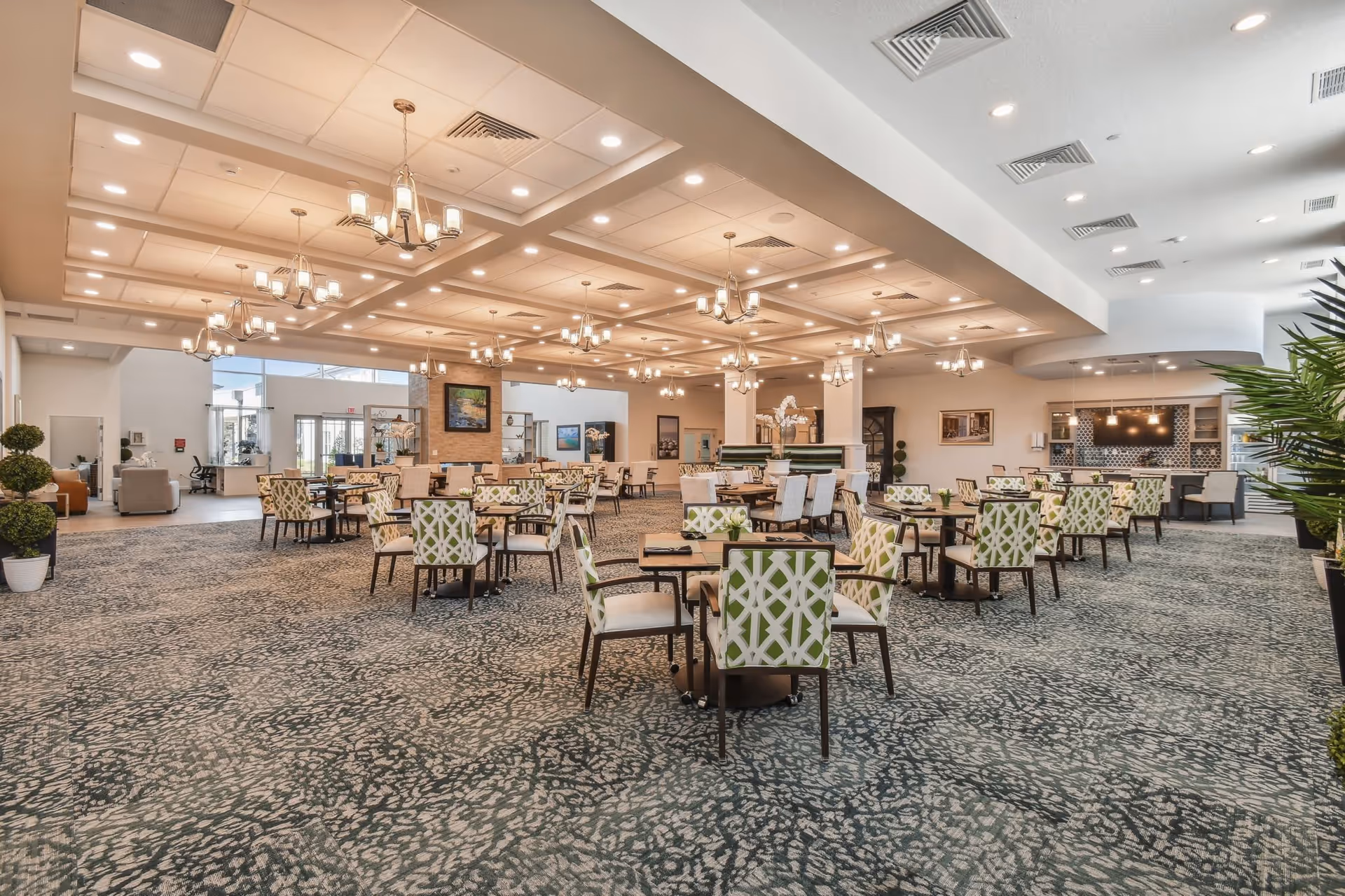 Spacious dining room with multiple tables and chairs featuring green and white patterned upholstery. The room has a patterned carpet, several chandeliers hanging from a coffered ceiling with recessed lighting, and large windows allowing natural light. There are plants and decorative elements around the room, and a counter area with pendant lights in the background.