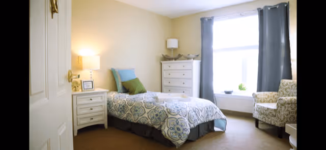 A bright and cozy bedroom with a single bed covered in a patterned blue and white bedspread, a white nightstand with a lamp and framed photo, a white chest of drawers with a lamp and decorative items, a large window with gray curtains, and a patterned armchair near the window.