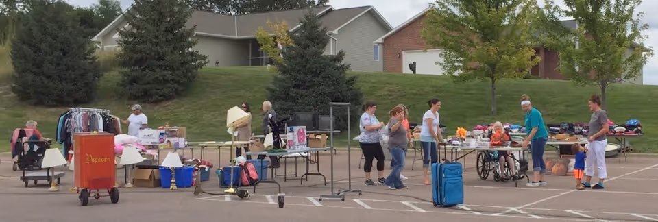 An outdoor community event in a parking lot with several people standing and interacting around tables filled with various items including clothes, lamps, and household goods. One person is seated in a wheelchair, and there are houses and trees in the background.