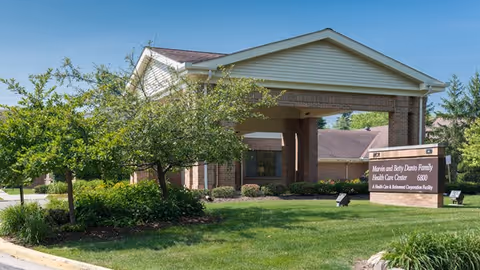 Exterior view of the Marvin and Betty Danto Family Health Care Center building with a covered entrance, surrounded by green grass, bushes, and trees under a clear blue sky.