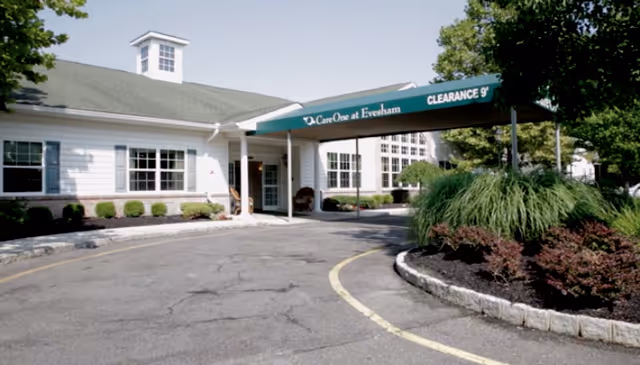 Exterior view of CareOne at Evesham facility showing a white building with green roof and a covered entrance with a green awning that reads 'CareOne at Evesham CLEARANCE 9'. There are landscaped bushes and trees around the driveway.