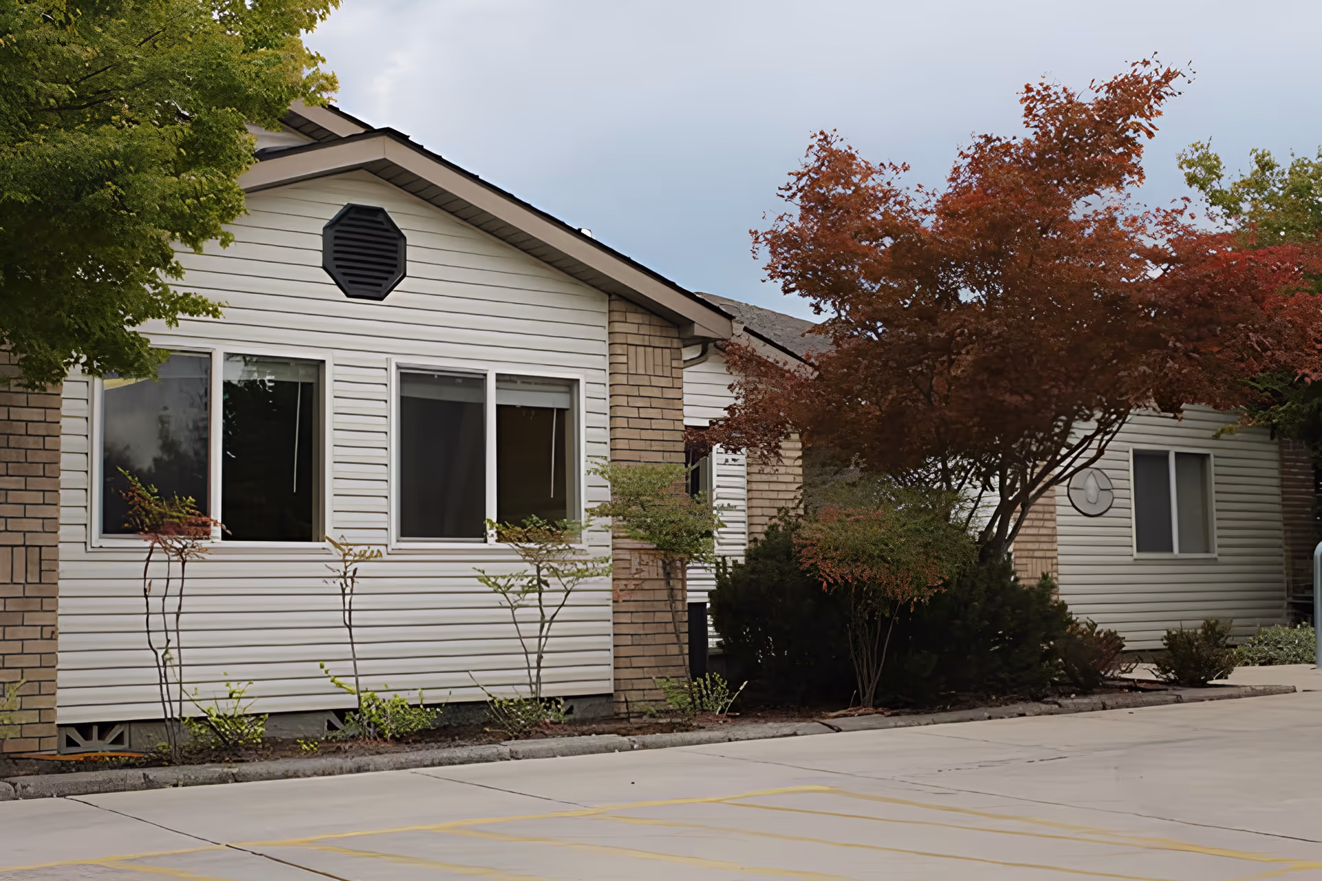 Exterior view of a single-story building with white siding and brick accents, surrounded by green and red-leaved trees and shrubs. The building has several windows and a paved area in front.