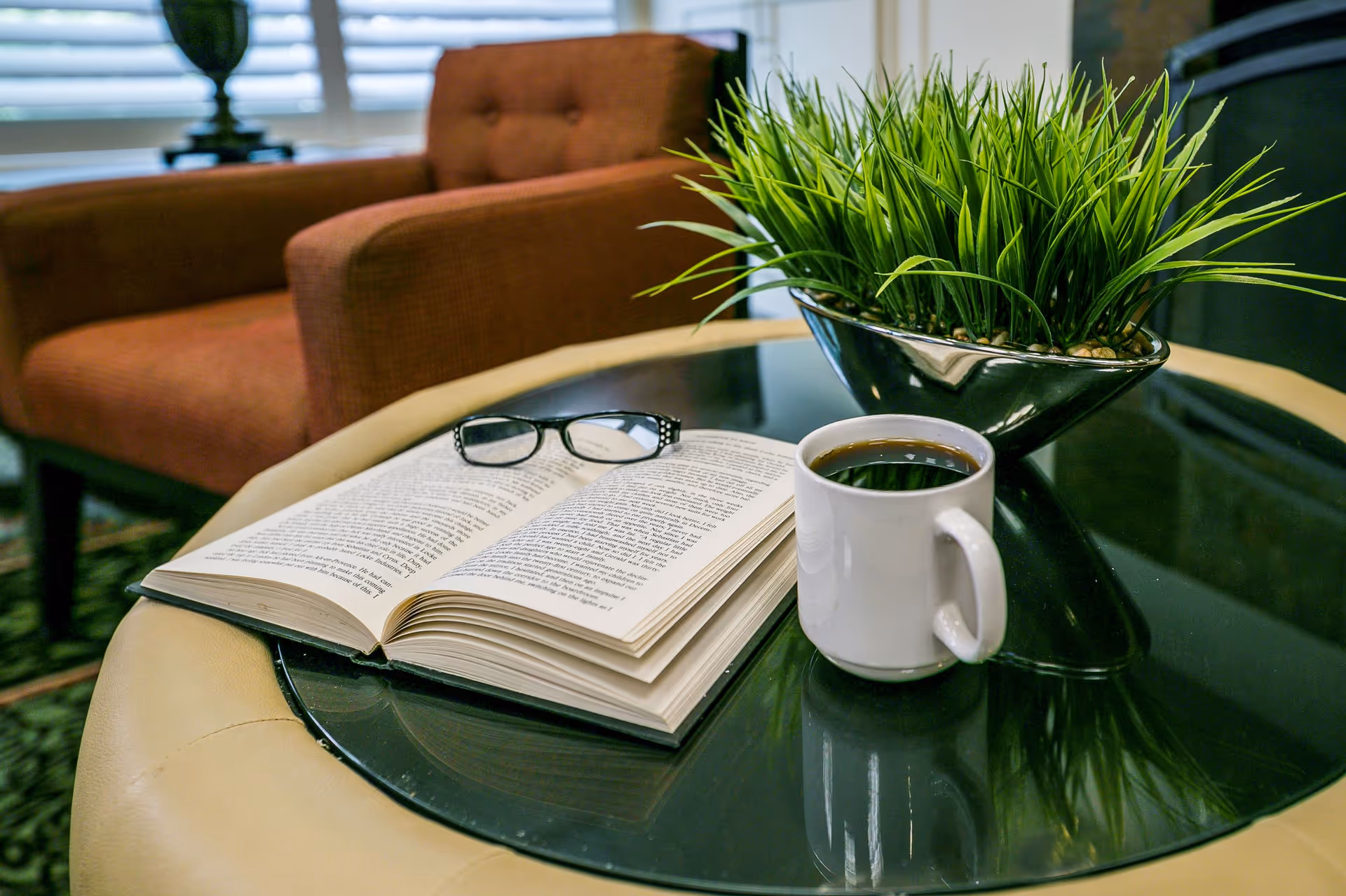 A cozy living room setting with a round glass-top table holding an open book with reading glasses on top, a white coffee mug filled with black coffee, and a decorative green plant in a silver bowl. In the background, there is a brown upholstered armchair and a window with white blinds.