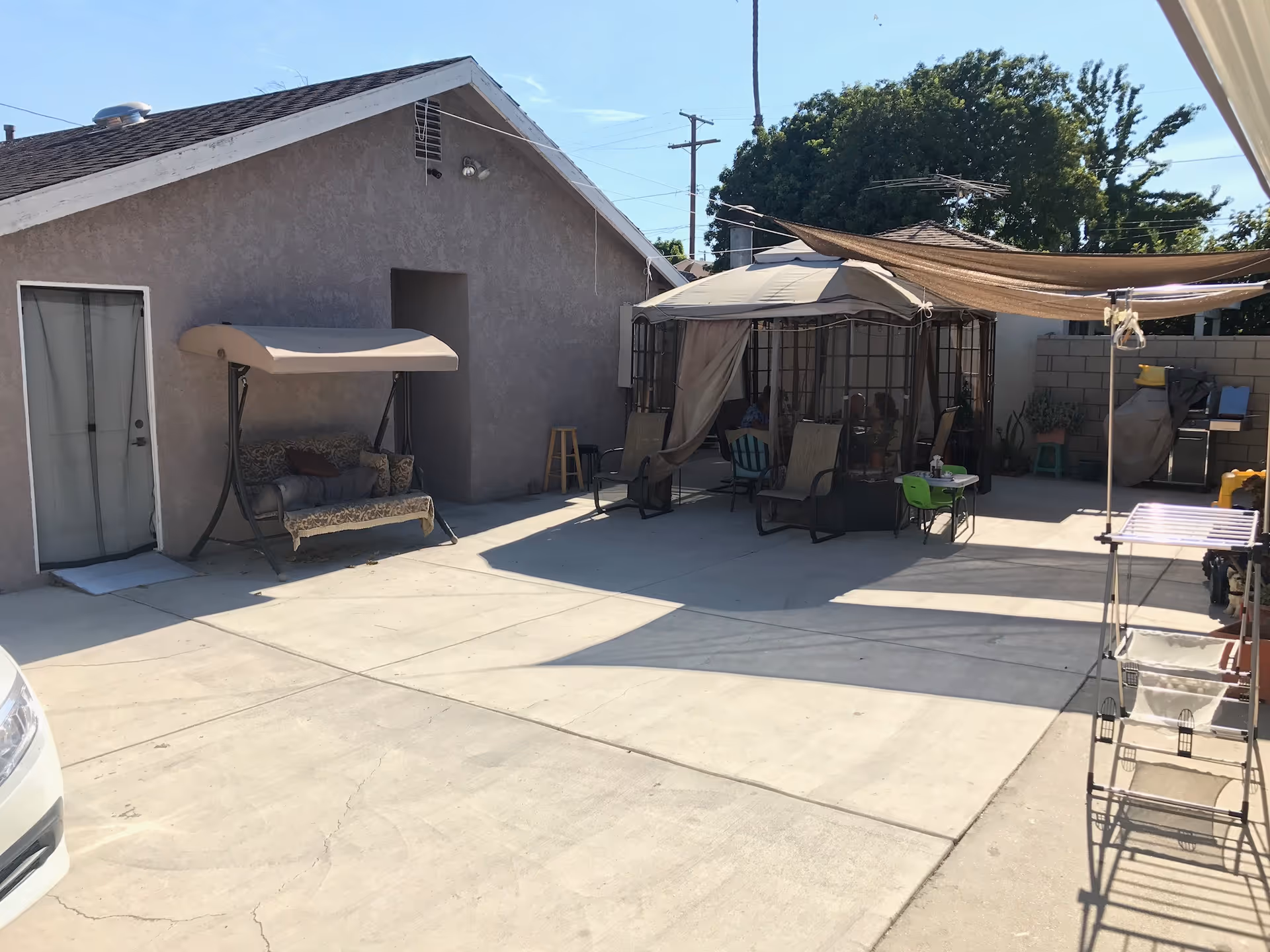 Outdoor patio area with a swing bench, a gazebo with chairs inside, a small table with a green chair, and a shaded canopy. The area is paved with concrete and surrounded by a block wall and a building with a door and a window.