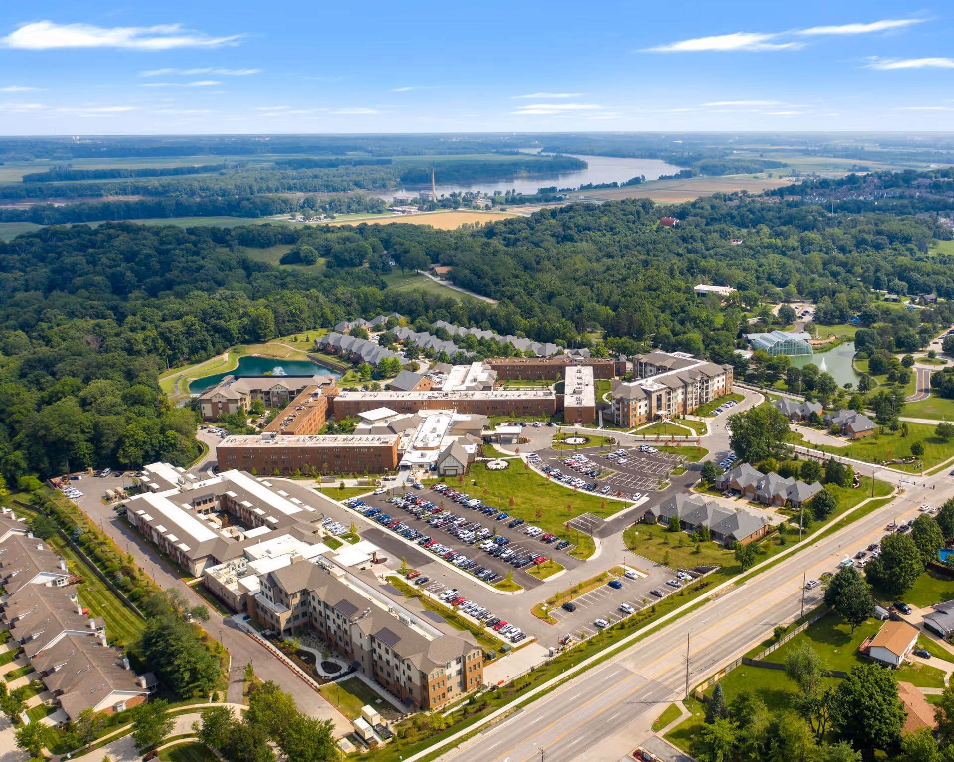 Aerial view of Friendship Village Chesterfield senior living facility showing multiple buildings, parking lots filled with cars, green lawns, trees, a pond with a fountain, and surrounding wooded and rural landscape under a partly cloudy sky.