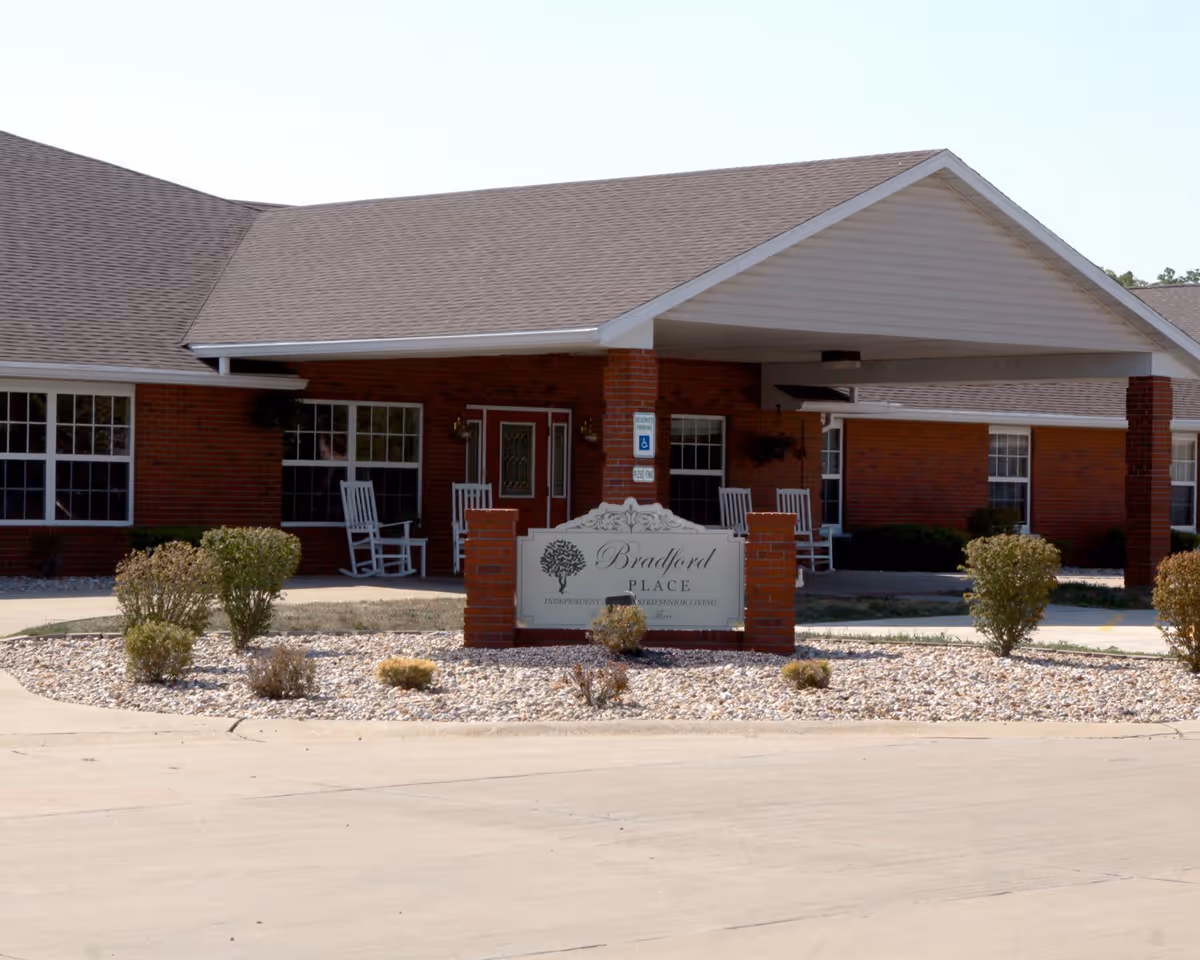 Exterior view of Bradford Place, a single-story brick building with a covered entrance. There are white rocking chairs on the porch and a landscaped area with small bushes and rocks in front of the building. A sign in front reads 'Bradford Place'.