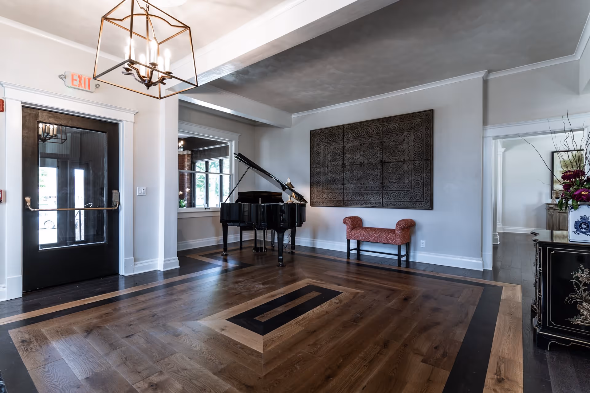 Interior view of a room with a grand piano, a red upholstered bench, decorative wall art, and a black door with an exit sign above it. The room has wooden flooring with a decorative pattern and a modern chandelier hanging from the ceiling.