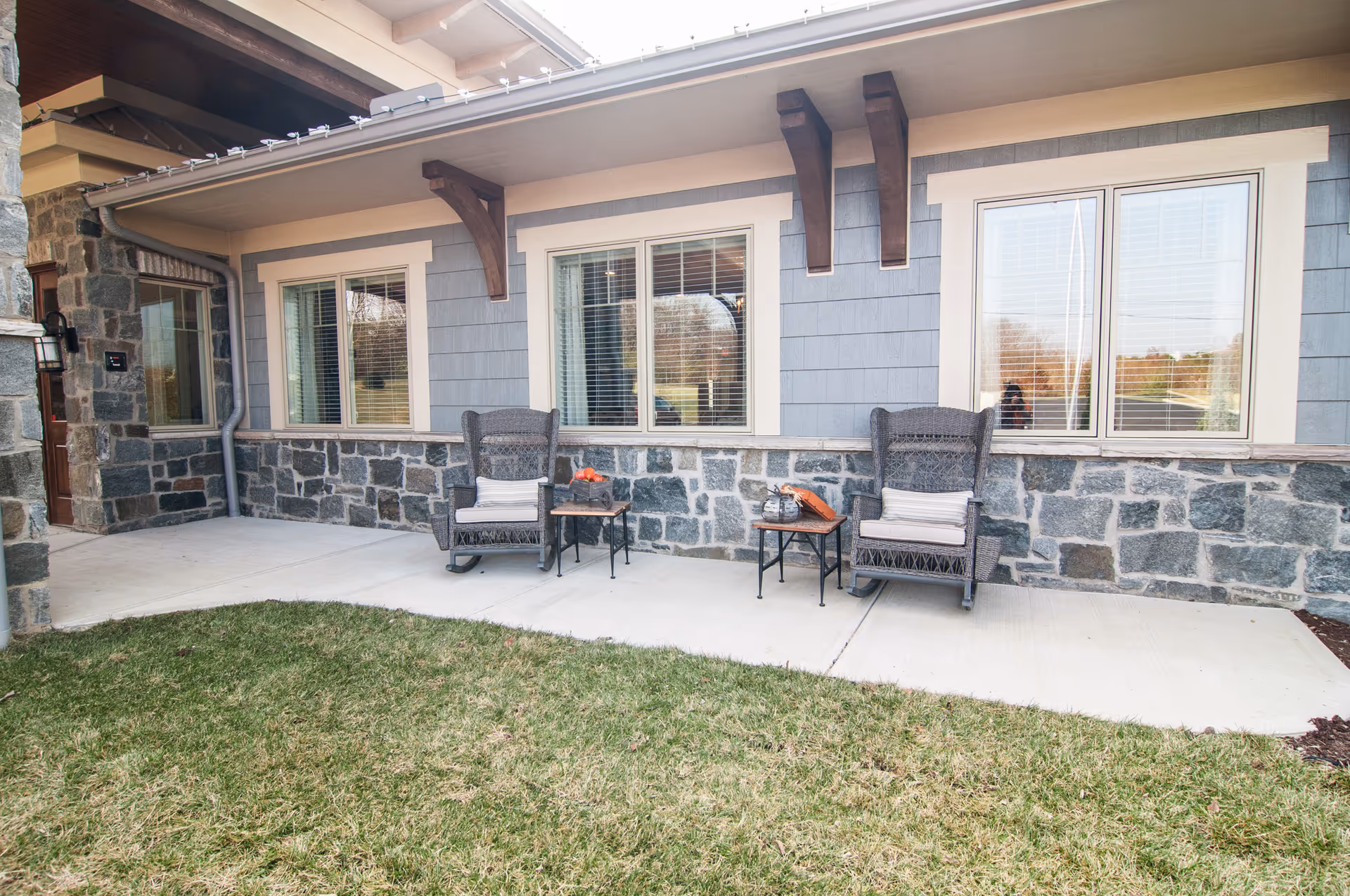Outdoor patio area of a senior living facility with two wicker rocking chairs, each with a cushion, and two small side tables placed between and beside the chairs. The building exterior features stone and blue siding with three windows and wooden beams supporting the roof overhang. There is a grassy lawn in front of the patio.