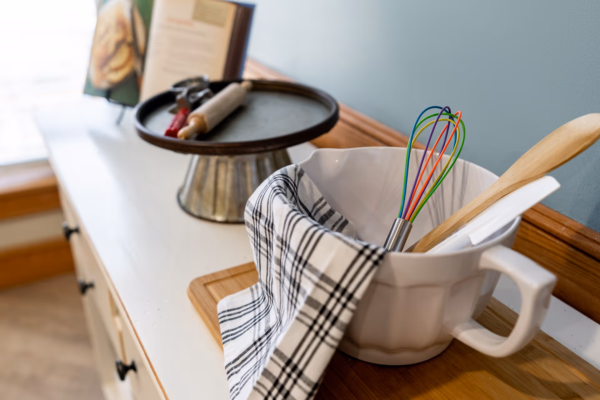 A white mixing bowl on a wooden cutting board with a black and white checkered kitchen towel draped over the side. Inside the bowl are a colorful whisk, a wooden spoon, and a white spatula. In the background, there is a metal cake stand with a rolling pin and cookie cutters on top, and a blurred menu or recipe card.