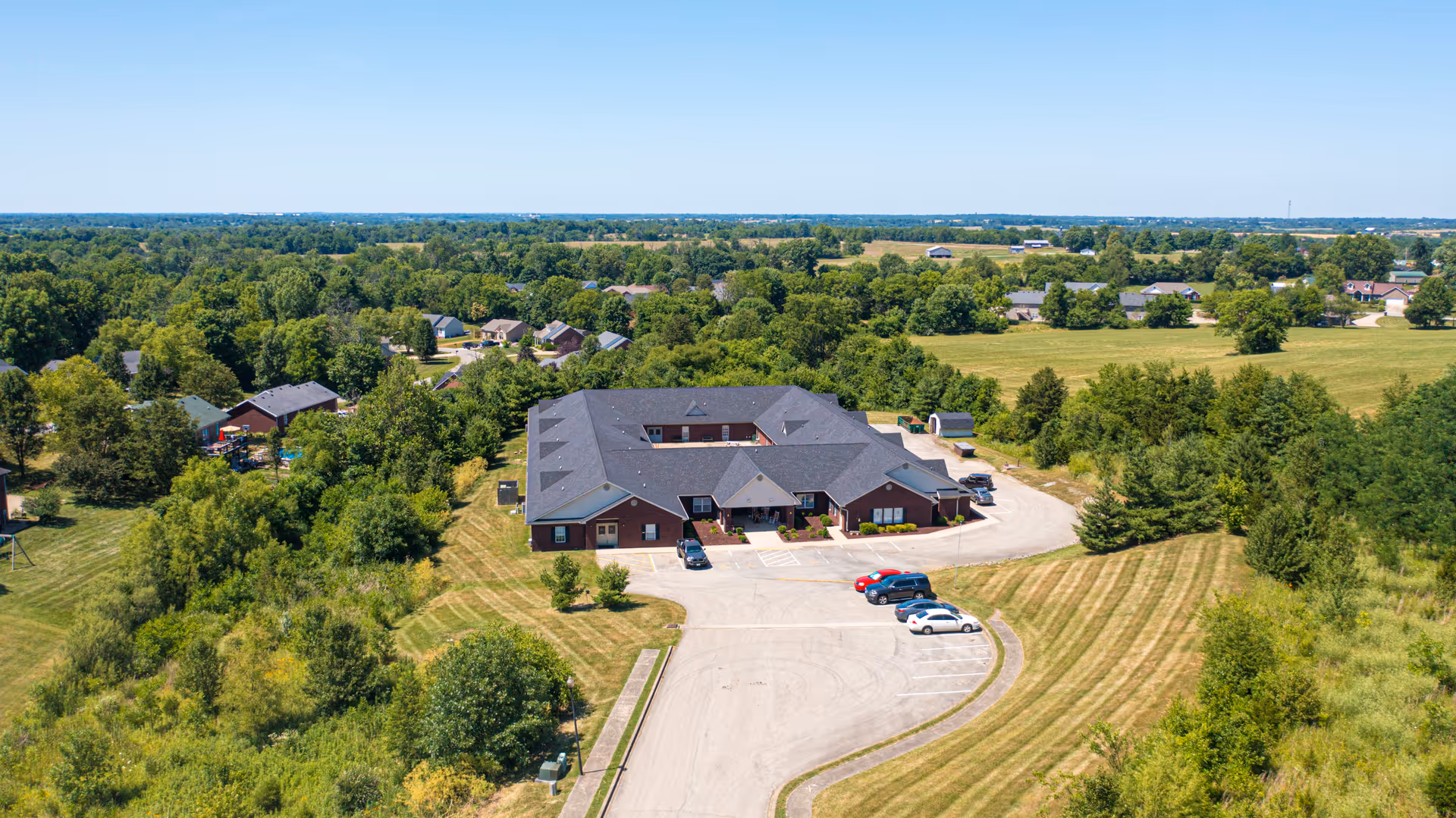 Aerial view of a single-story brick building with a dark roof, surrounded by green trees and open grassy areas. There is a parking lot in front of the building with a few cars parked. The landscape extends into the distance with more trees and scattered houses under a clear blue sky.