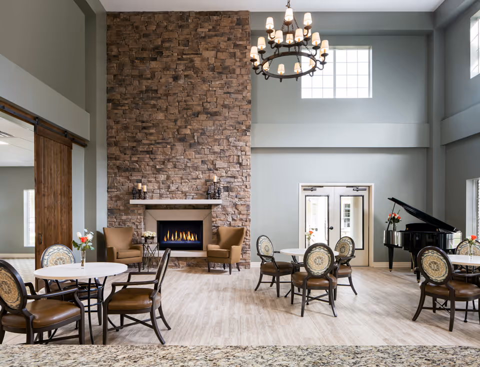 A spacious and elegant common area with a tall stone fireplace, two brown armchairs, round tables with chairs, a grand piano, and large windows letting in natural light. A chandelier hangs from the high ceiling.