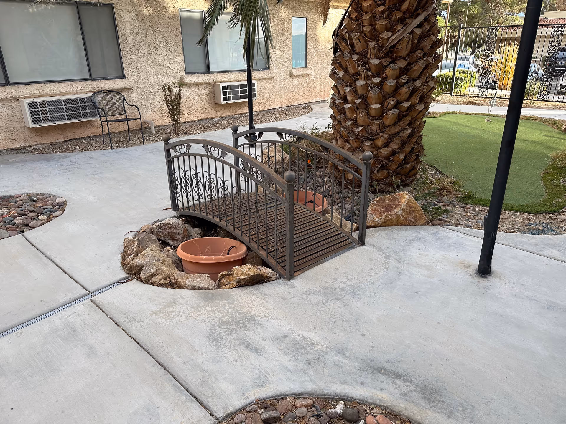 Outdoor area with a small decorative wooden bridge over a dry rock bed containing a large empty flower pot. There is a large palm tree next to the bridge, concrete walkways, a metal chair against a beige stucco building wall with windows and air conditioning units, and a fenced area with green artificial turf in the background.