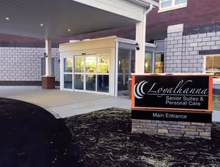 Entrance to Loyalhanna Senior Suites and Personal Care facility showing automatic sliding glass doors under a covered drop-off area, with a sign in front that reads 'Loyalhanna Senior Suites & Personal Care Main Entrance'.
