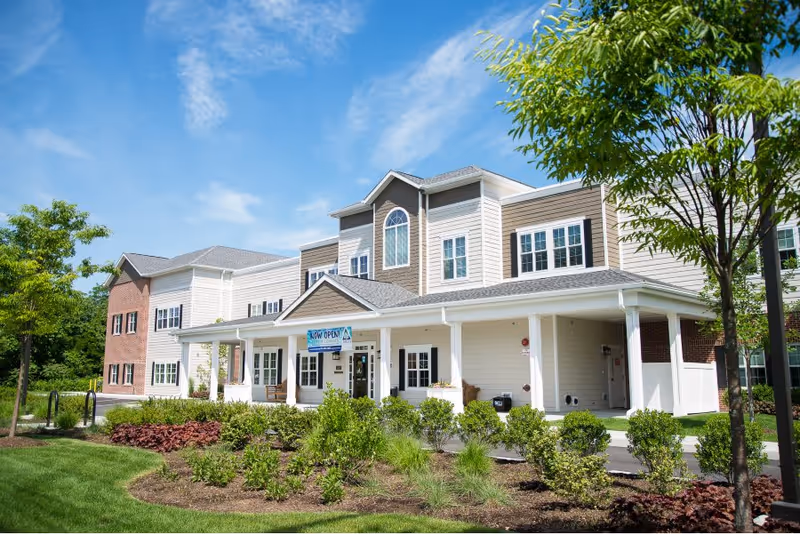 Exterior view of a two-story senior living facility named Artis with a covered entrance, well-maintained landscaping including bushes and trees, and a clear blue sky.