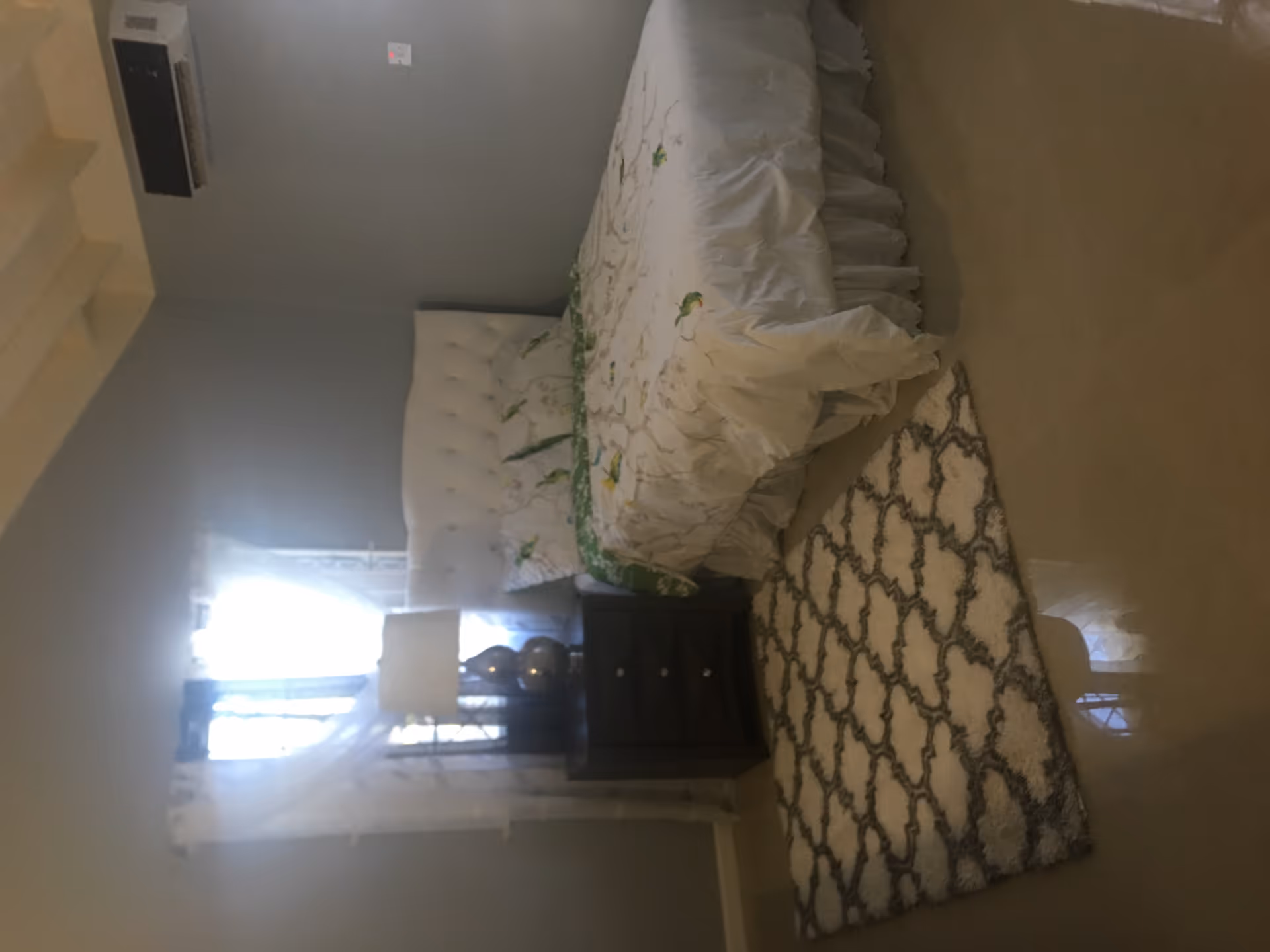A bedroom with a white tufted headboard bed covered with a white bedspread featuring a floral pattern. Next to the bed is a dark wooden nightstand with a lamp and decorative items. A patterned rug is placed on the floor beside the bed. Light filters in through a window with sheer curtains.
